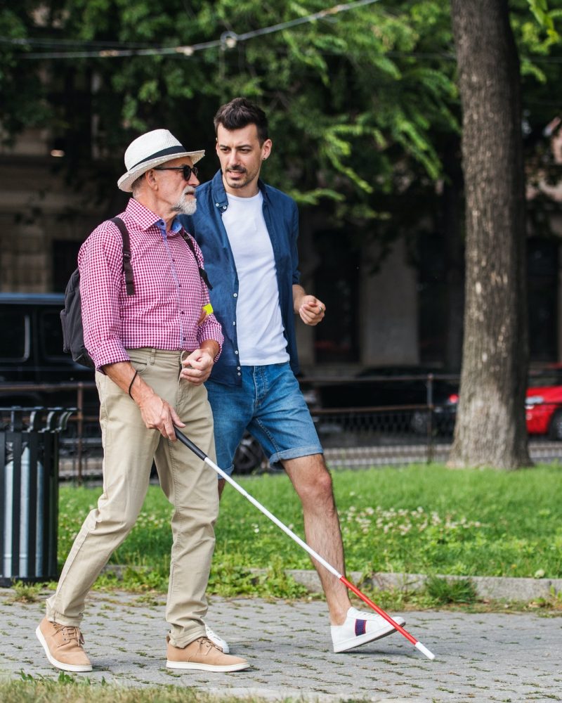 Young man and blind senior with white cane walking on pavement in city